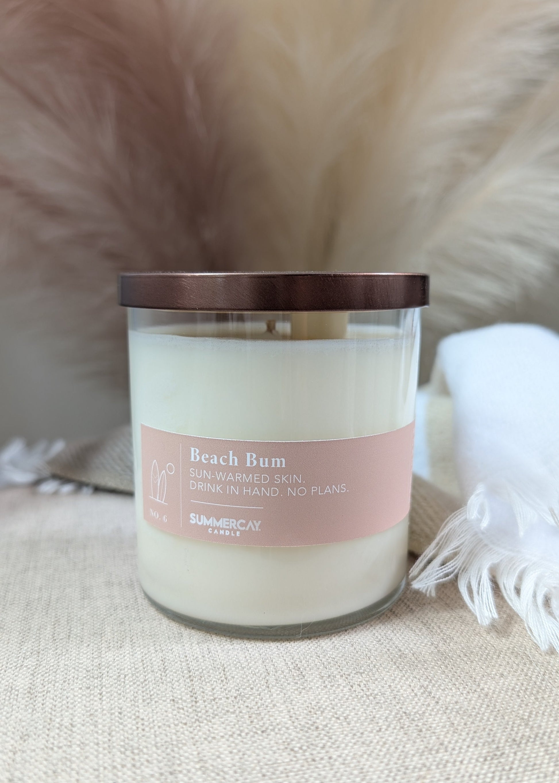 A candle in a clear glass jar with a label reading 'Beach Bum' on a beige textured surface with a fluffy cream background.