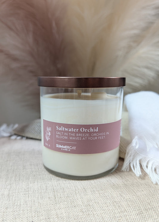 A jar candle labeled 'Saltwater Orchid' in a clear glass jar with a bronze lid, on a soft beige fabric background with fluffy decorative elements.
