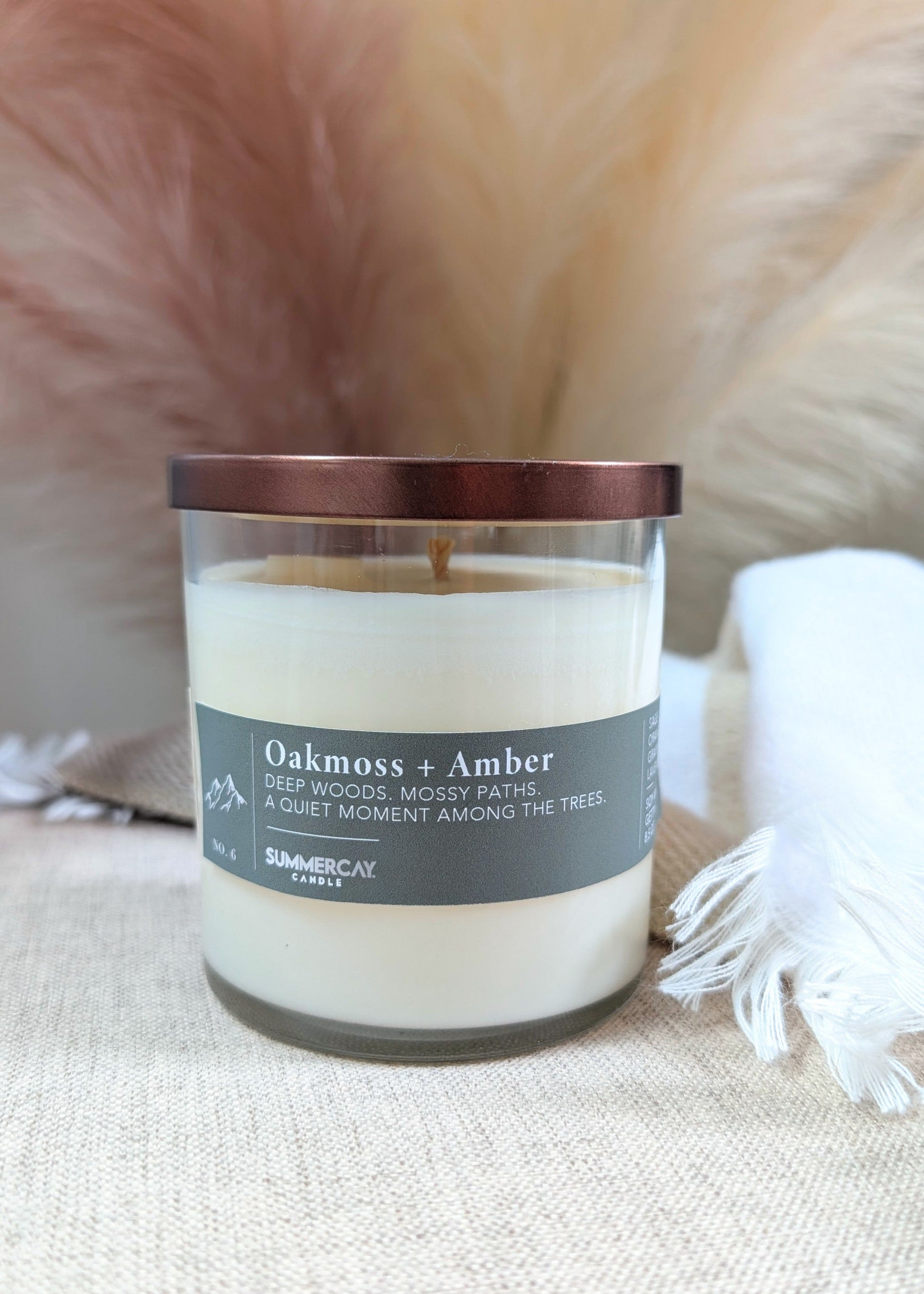 A glass jar candle with a bronze lid, labeled 'Oakmoss + Amber' on a beige fabric surface with soft neutral background props.
