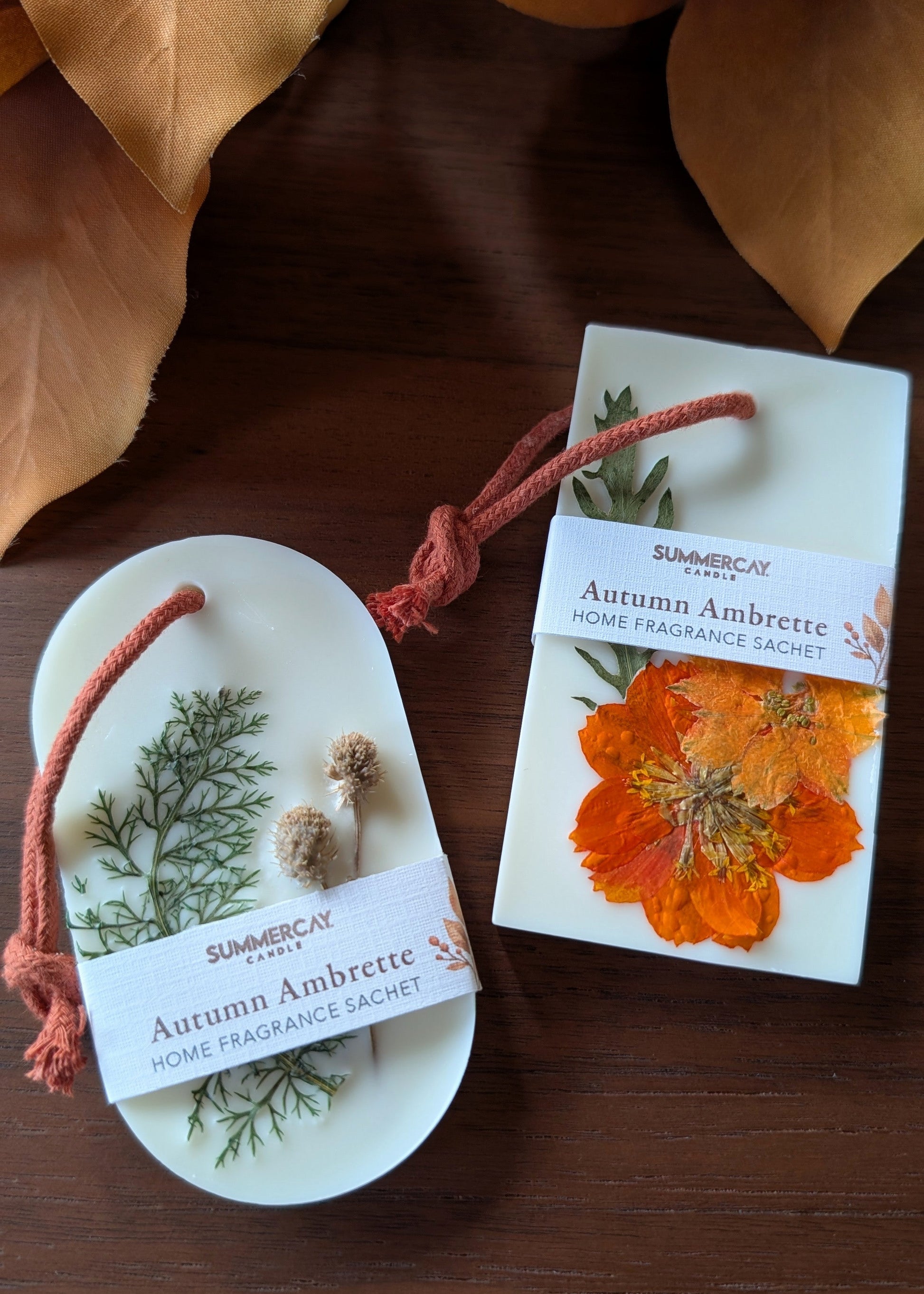 Two SummerCay Scents 'Autumn Ambrette' home fragrance sachets displayed on a dark wooden surface with dried autumn leaves. Shown unwrapped, revealing a white wax base with a pressed orange flower and green botanical elements tied with a rust cord.