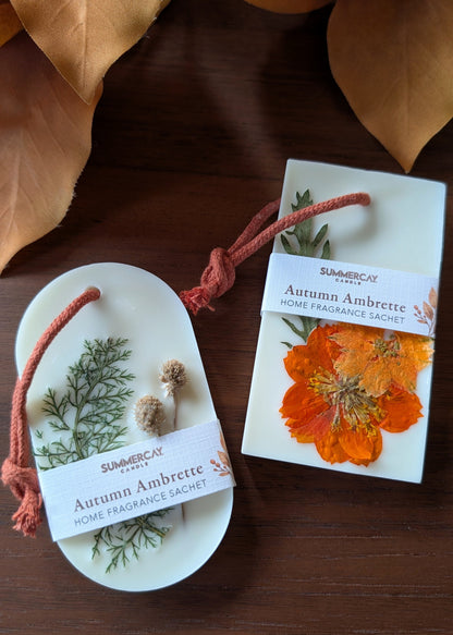 Two SummerCay Scents 'Autumn Ambrette' home fragrance sachets displayed on a dark wooden surface with dried autumn leaves. Shown unwrapped, revealing a white wax base with a pressed orange flower and green botanical elements tied with a rust cord.