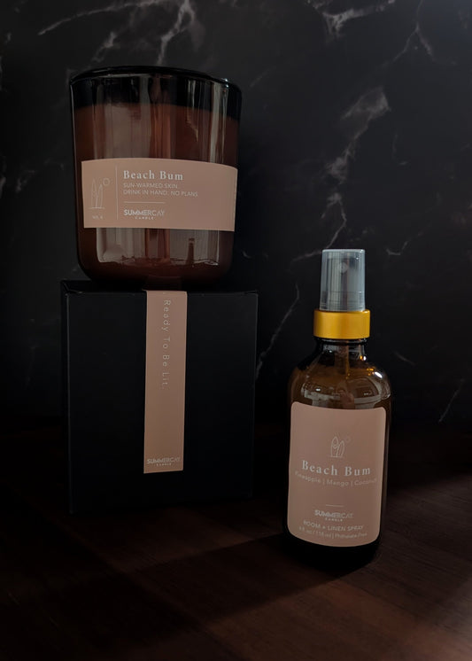 Beach bum amber glass candle on top of gift box with matching room + linen spray on a wooden table.