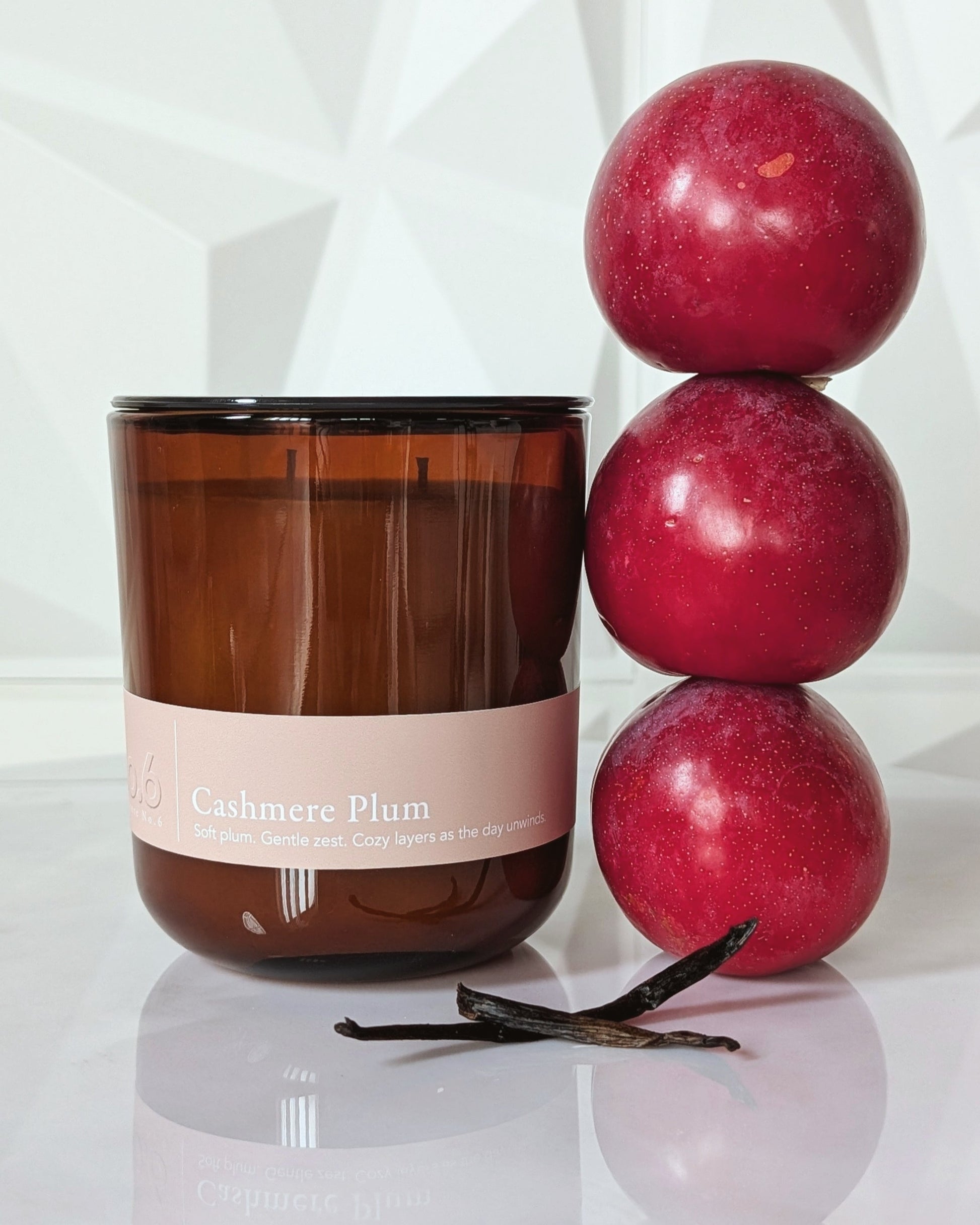 Candle labeled 'Cashmere Plum' with plums and vanilla beans on a reflective surface.