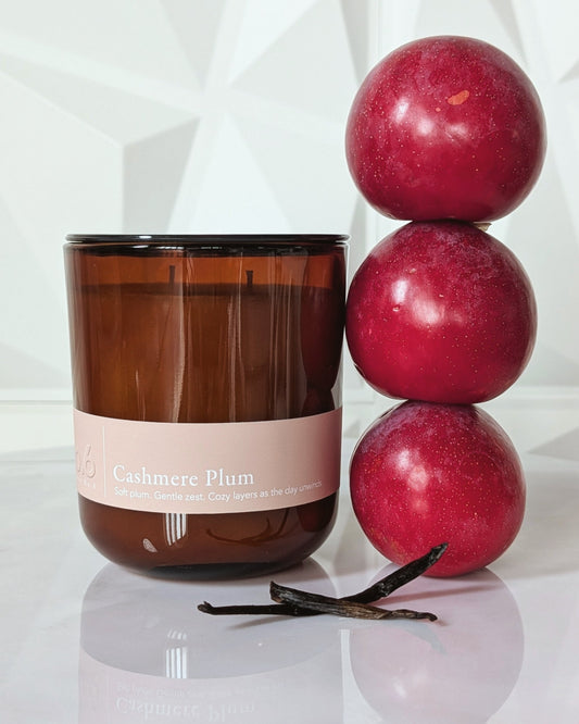 Candle labeled 'Cashmere Plum' with plums and vanilla beans on a reflective surface.