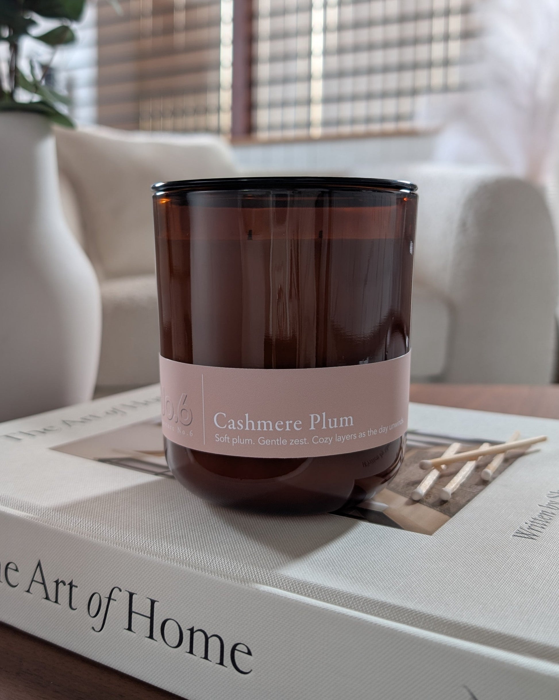 cashmere plum scented candle in an amber vessel on top of a book in a cozy indoor setting. 