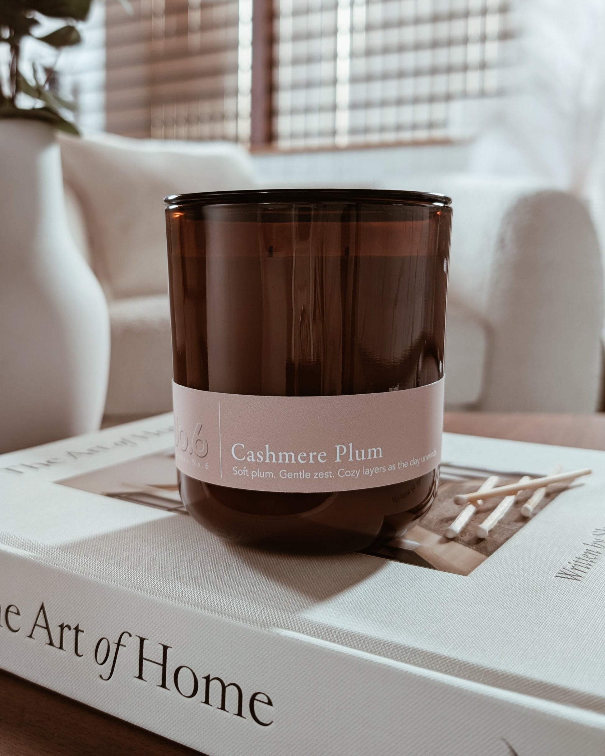 Amber glass candle with Cashmere Plum label on a book in a cozy indoor setting.