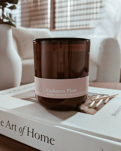 Amber glass candle with Cashmere Plum label on a book in a cozy indoor setting.