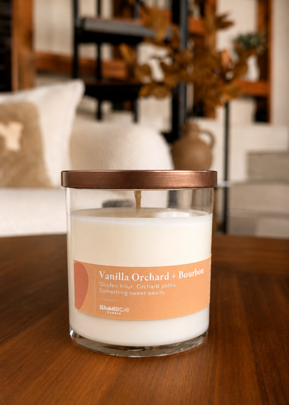 Vanilla orchard and bourbon scented candle in a clear glass vessel with bronze lid sitting on a wooden table with a blurred living room in the background.
