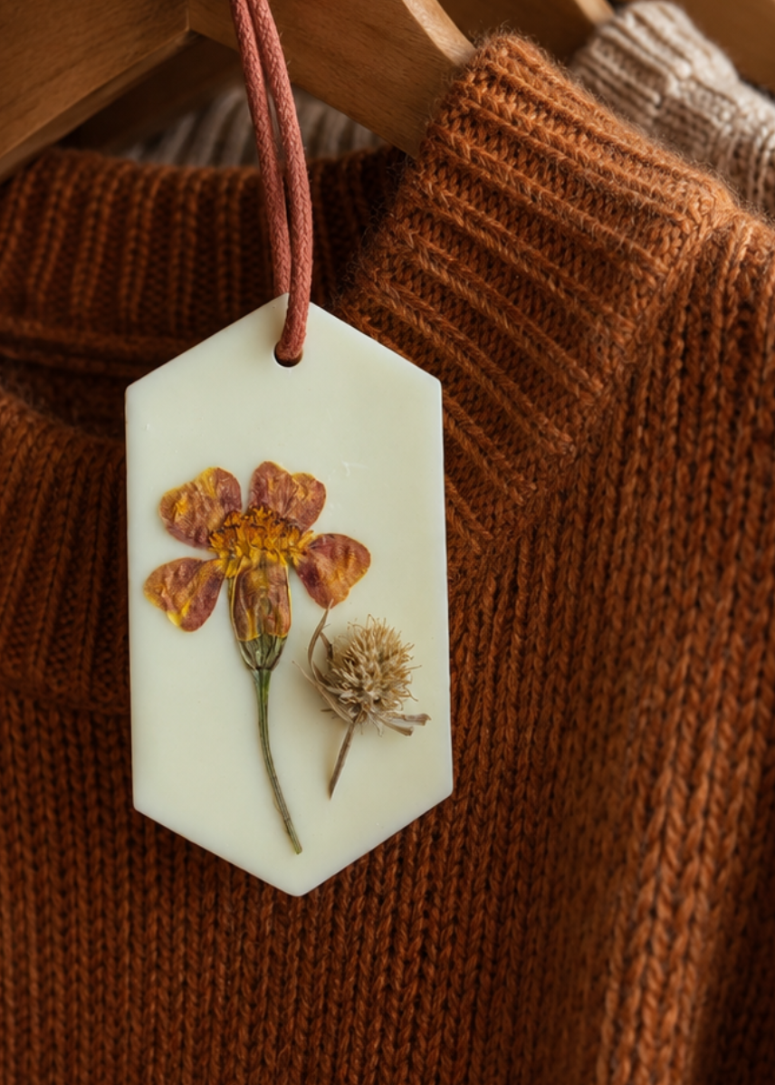 A close-up of a rust-brown knit sweater hanging on a wooden hanger from a clothing rack, with a small hexagonal white wax sachet featuring a pressed orange flower and dried seed pod hanging from a metal hook.
