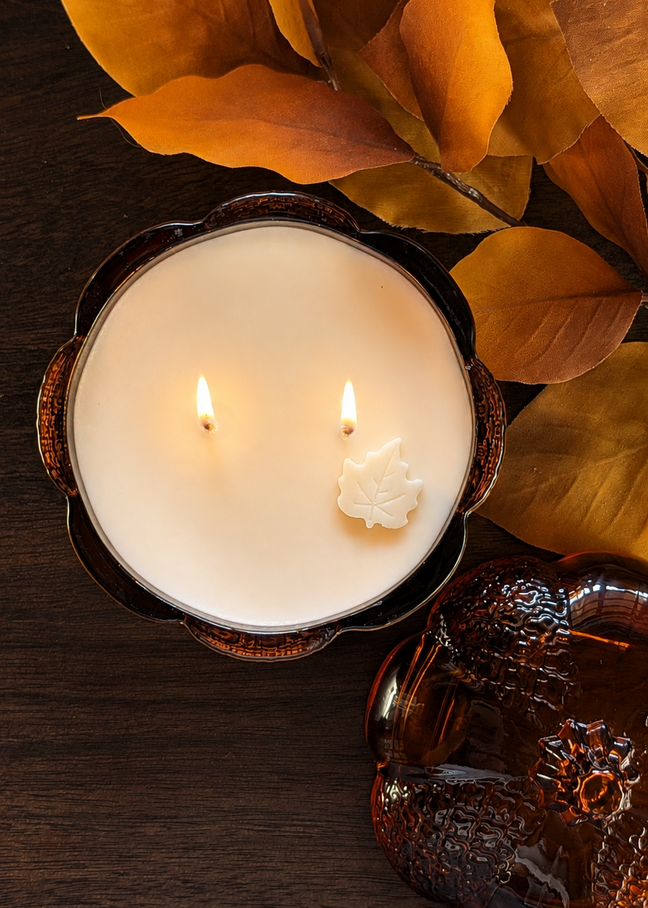 A lit double-wick candle in a dark amber pumpkin shaped glass, styled with autumn leaves on a dark wooden surface.