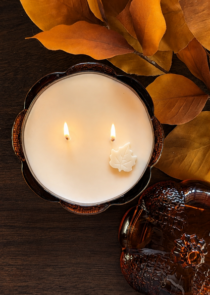A lit double-wick candle in a dark amber pumpkin shaped glass, styled with autumn leaves on a dark wooden surface.