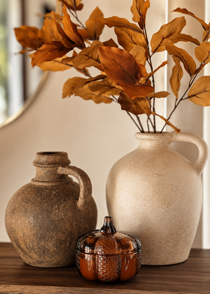 Two rustic decorative vases arranged on a wooden surface, with dried orange-brown leaves in the tallest cream vase, a smaller brown vase to the left, and a small amber glass pumpkin lidded candle in front.