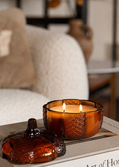 An amber glass pumpkin shaped jar candle with two lit wicks sits on a book in a softly styled indoor setting, with its matching amber lid placed beside it.