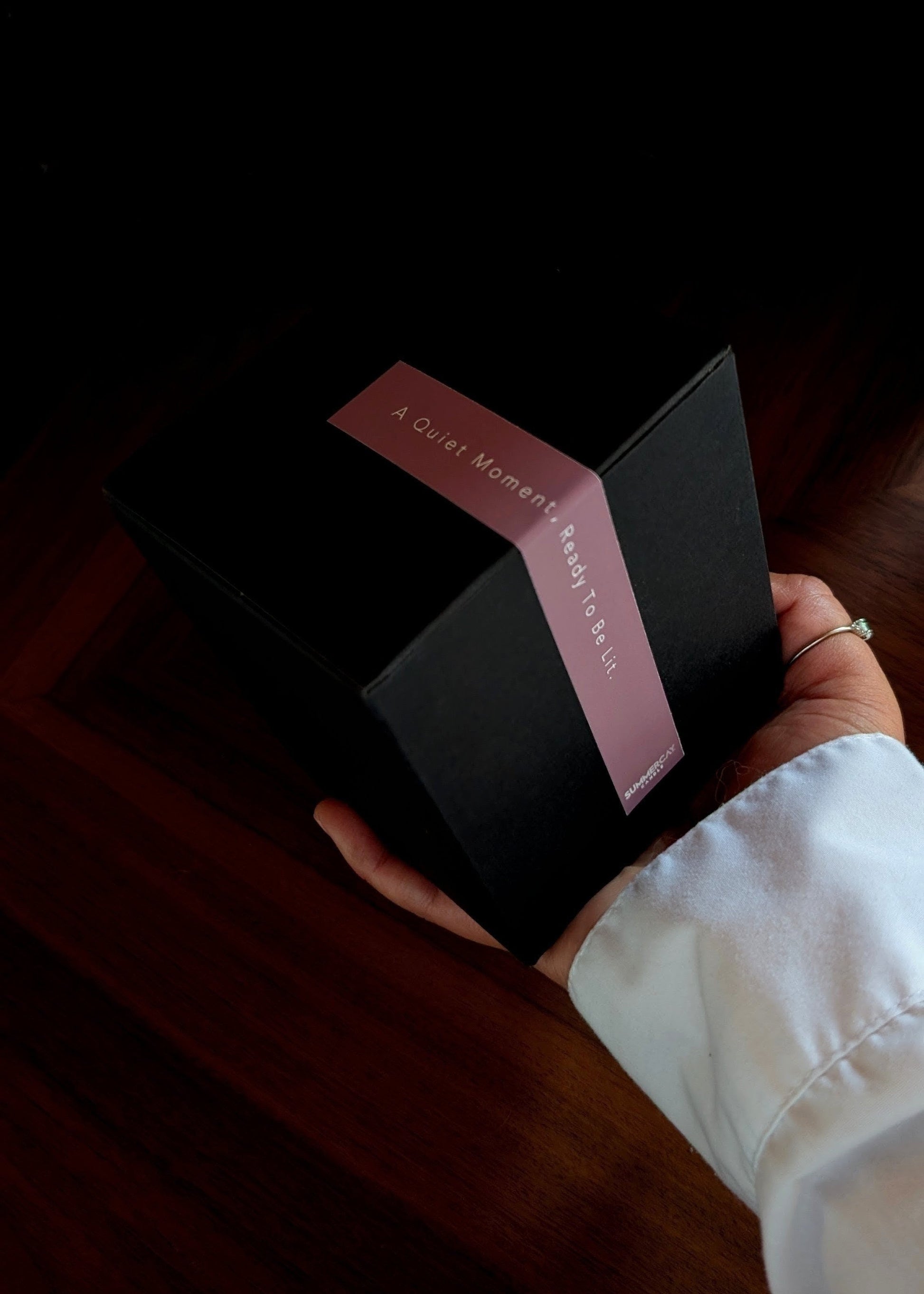 Hand holding a black box with a pink ribbon and text on a dark surface