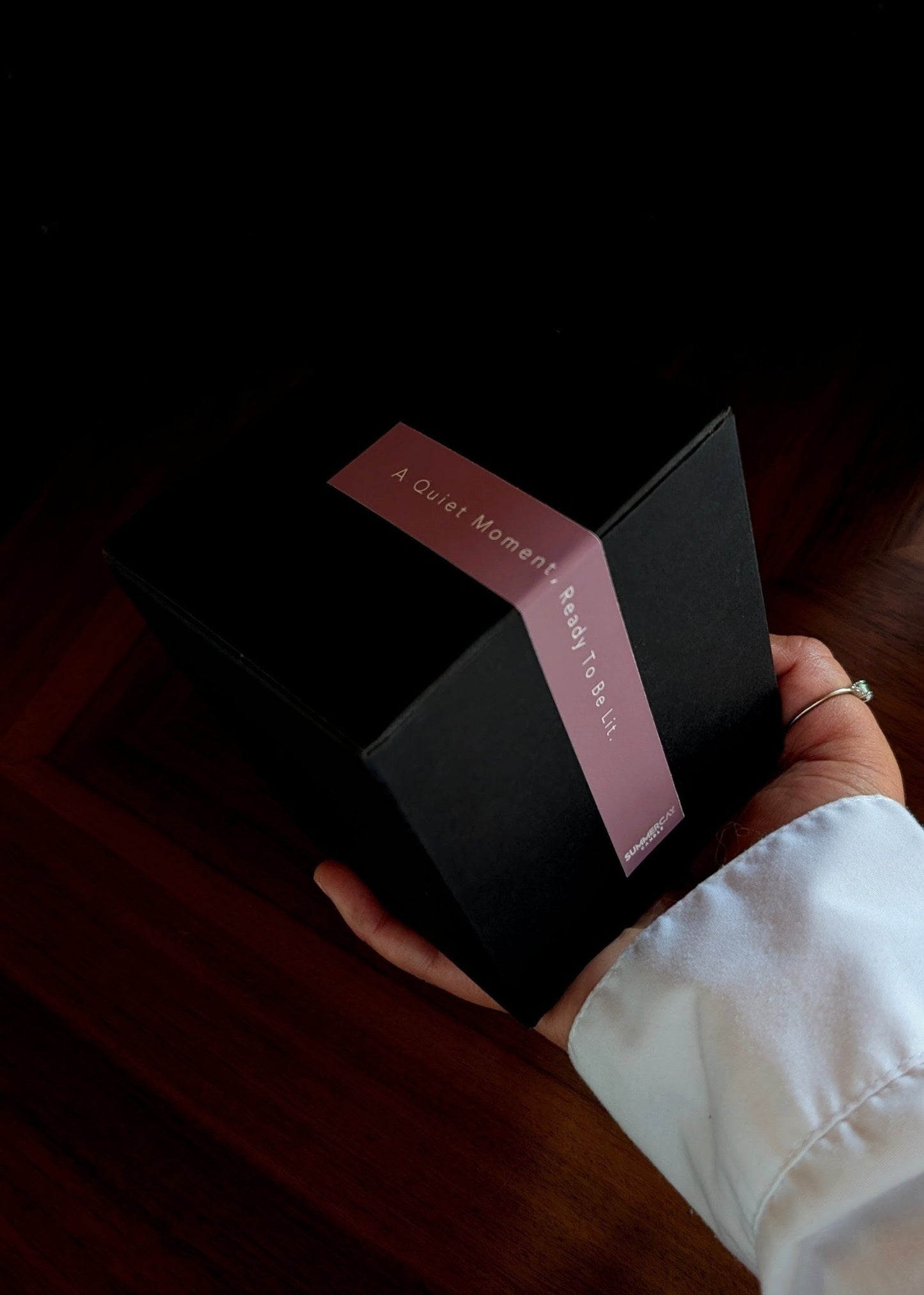 Hand holding a black box with a pink ribbon and text on a dark surface