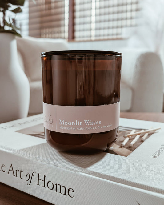 Amber glass candle labeled 'Moonlit Waves' on a book with a blurred cozy indoor setting.