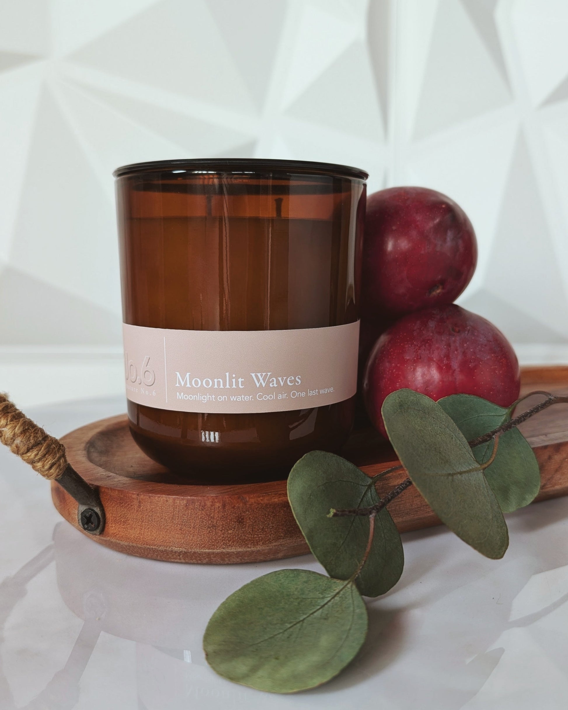 Candle with a label on a wooden tray with plums and eucalyptus leaves, against a white background