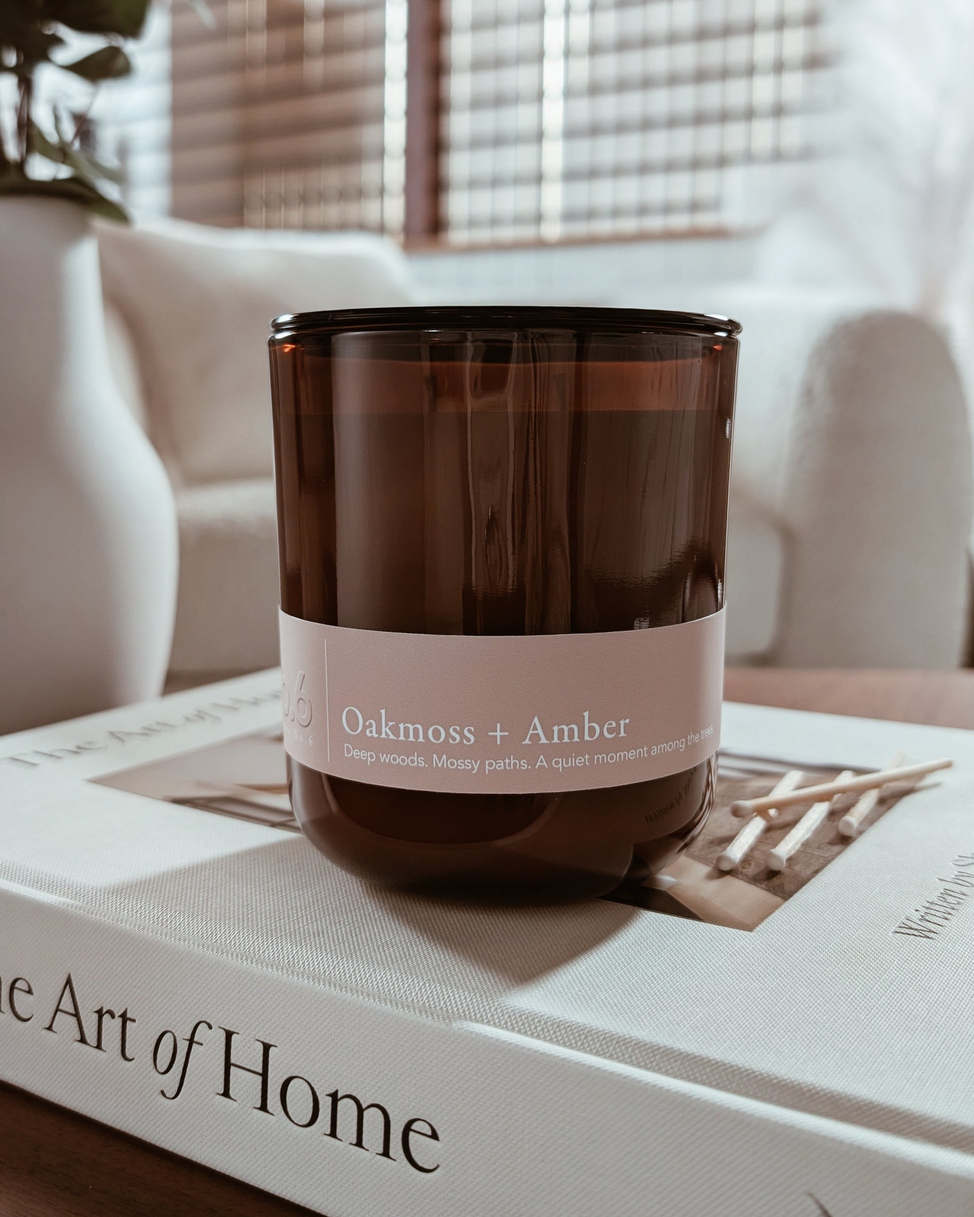 Amber glass candle labeled 'Oakmoss + Amber' on a book with a blurred cozy indoor setting.