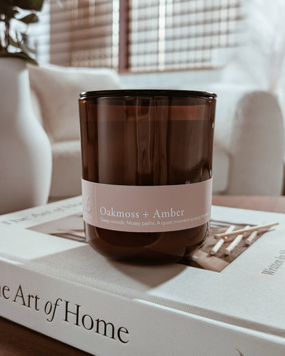 Amber glass candle labeled 'Oakmoss + Amber' on a book with a blurred cozy indoor setting.