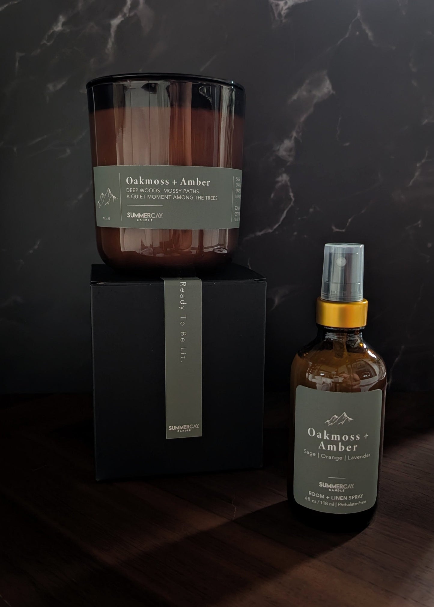 Oakmoss + Amber candle on a gift box with matching room + linen spray on a wooden table and dark background.