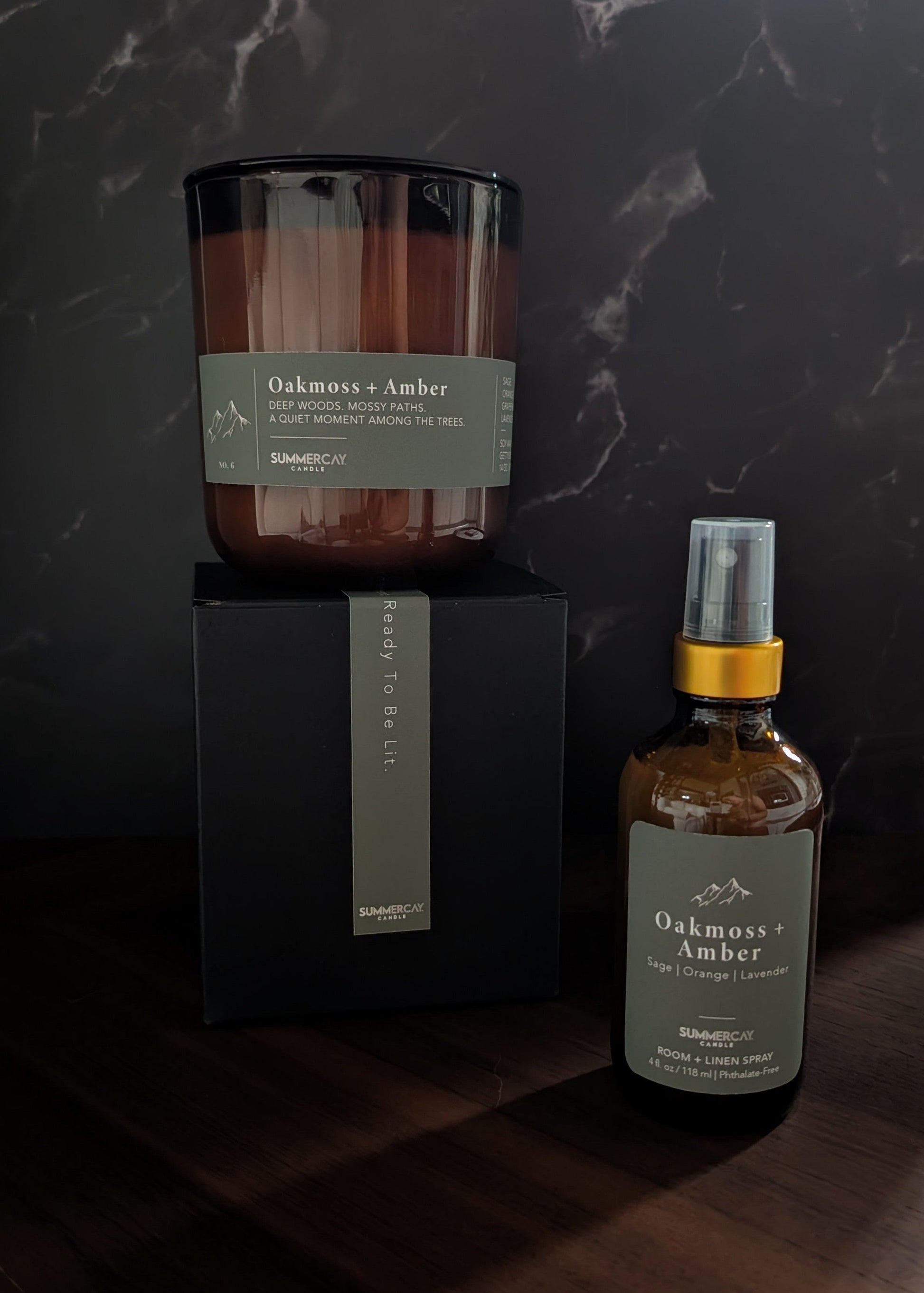 Oakmoss + Amber candle on a gift box with matching room + linen spray on a wooden table and dark background.