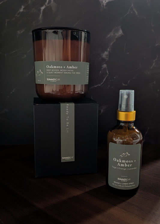 Oakmoss + Amber candle on a gift box with matching room + linen spray on a wooden table and dark background.