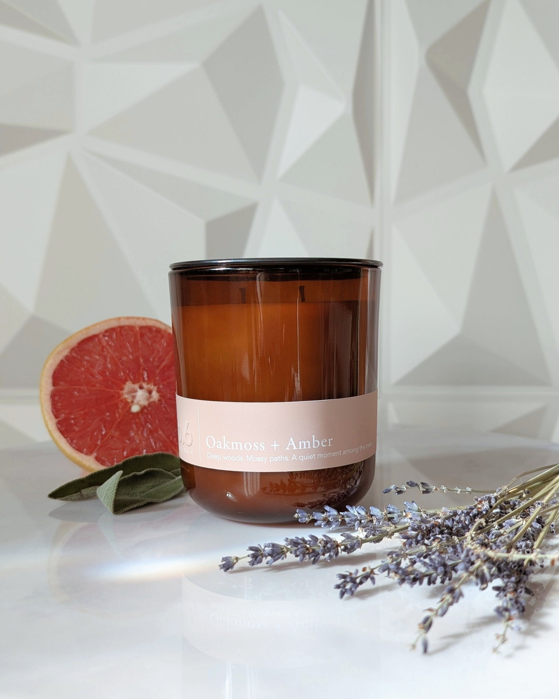 Candle with a Oakmoss + Amber label on a white surface with a grapefruit, sage, and lavender on a geometric wall background