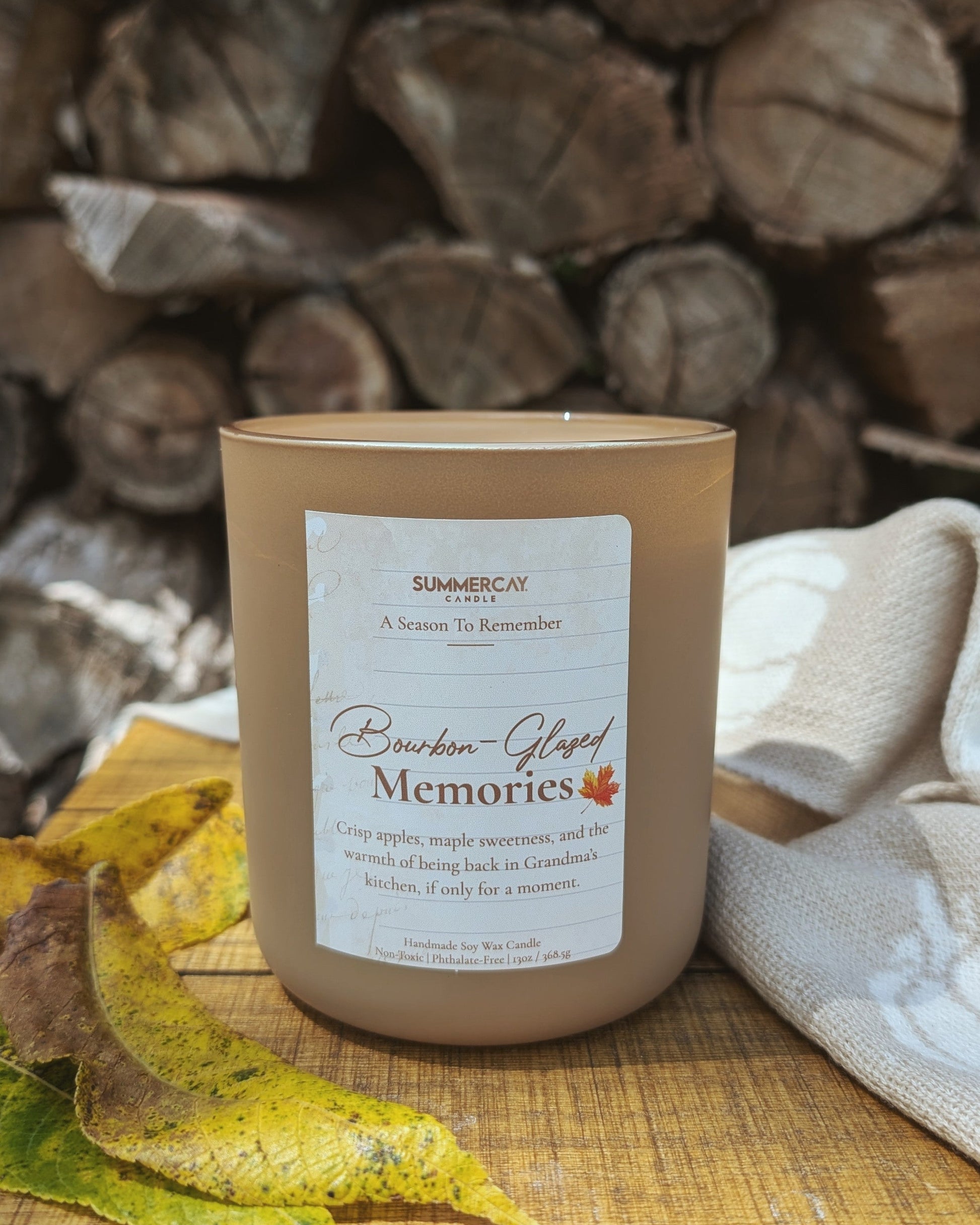 Candle labeled 'Bourbon Glazed Memories' on a wooden surface with stacked logs in the background
