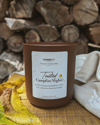 Candle labeled 'Toasted Campfire Nights' on a wooden surface with stacked logs in the background