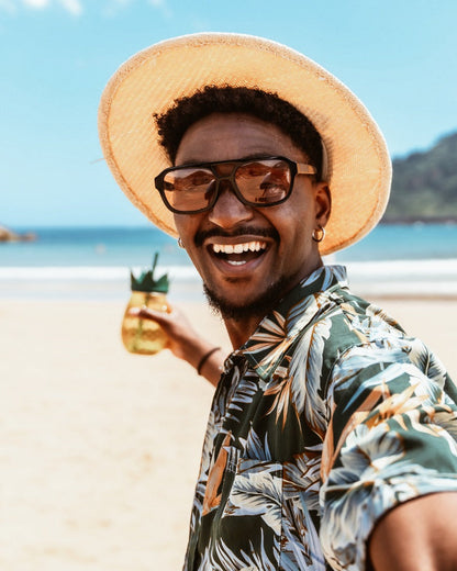 Man on a beach wearing a hat and sunglasses, holding a drink.