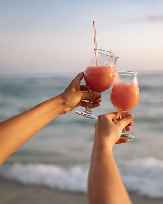 Two people holding glasses of pink drinks on a beach at sunset.