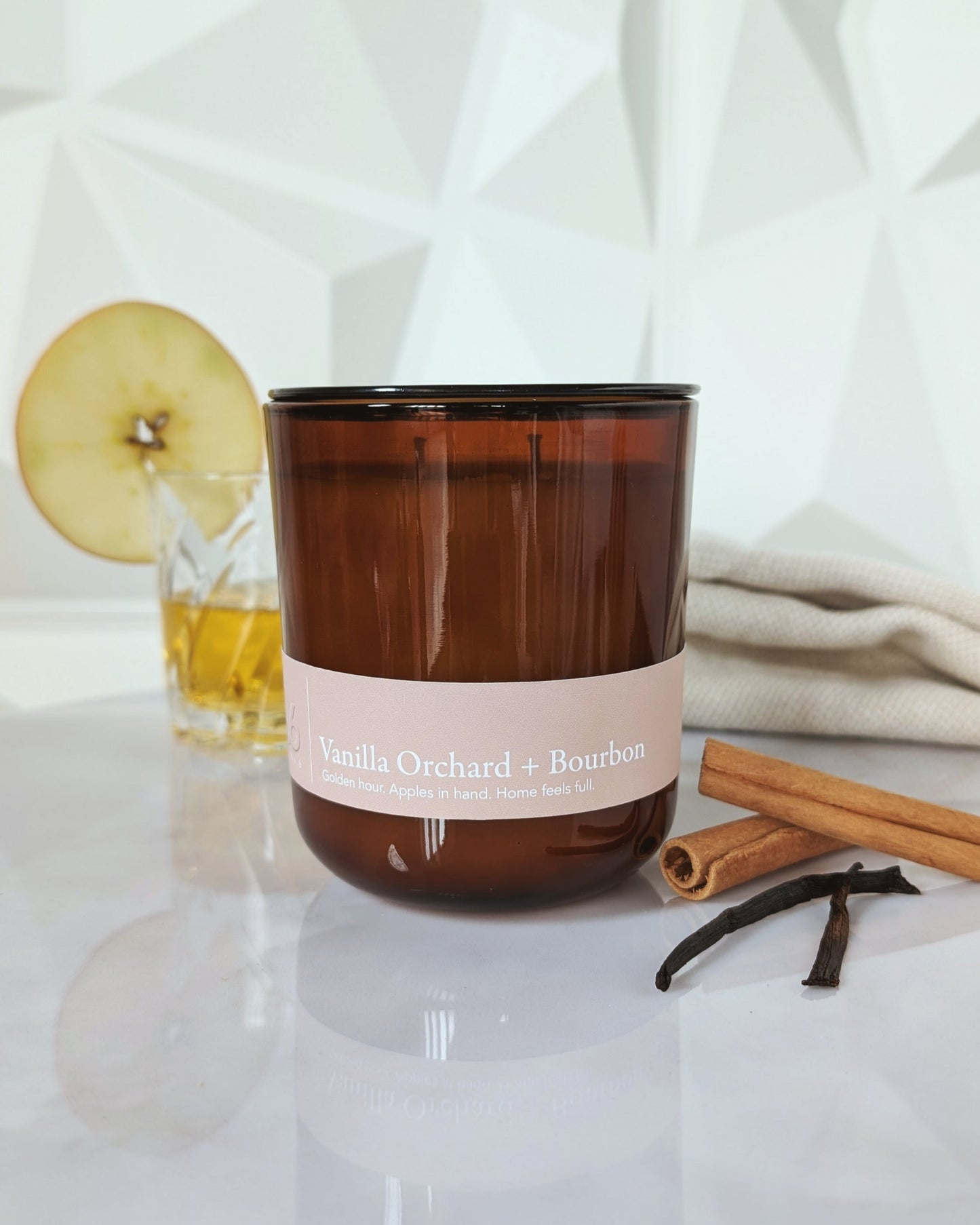 Vanilla Orchard + Bourbon candle with a drink and apple slice in the background, cinnamon and vanilla.