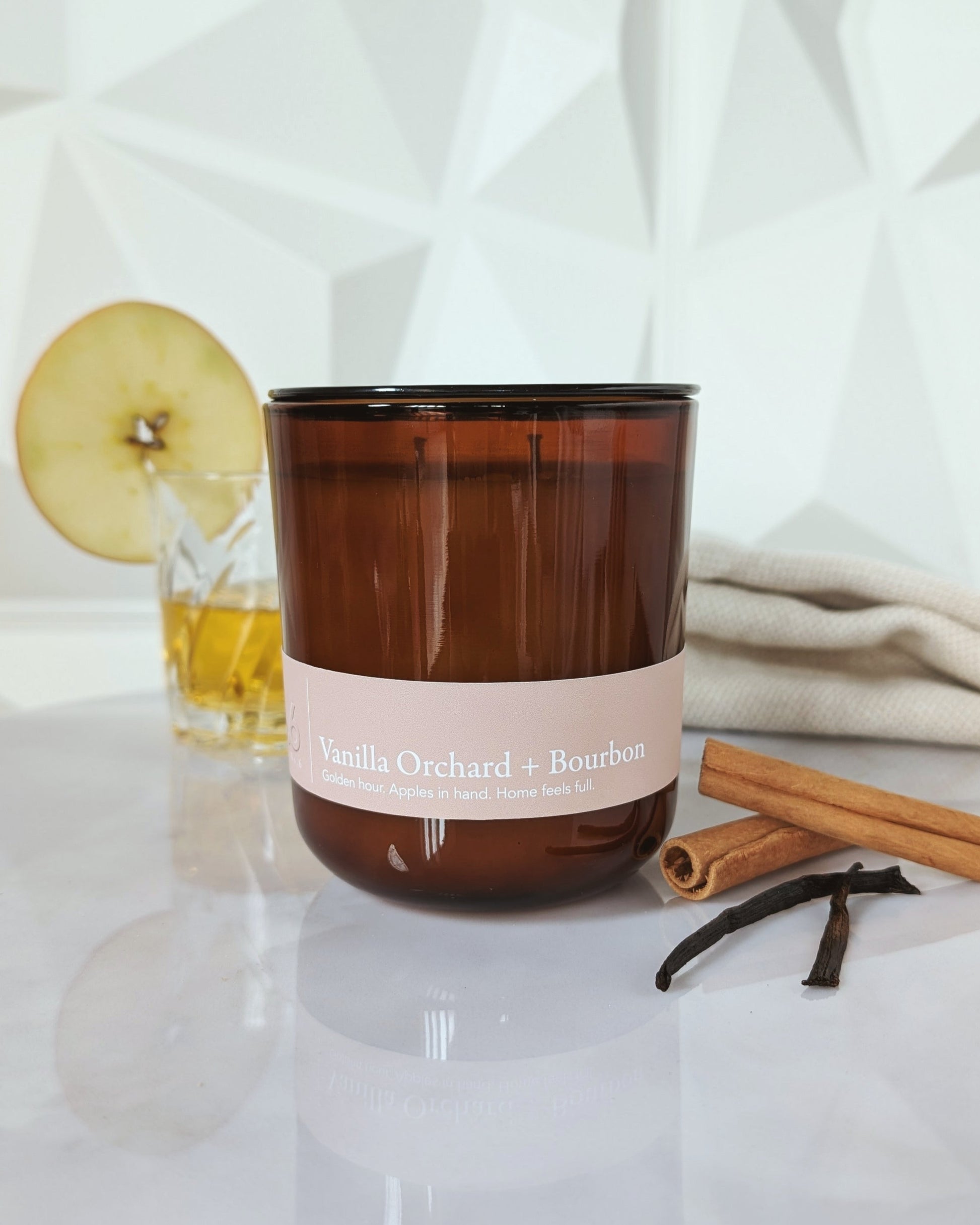 Vanilla Orchard + Bourbon candle with a drink and apple slice in the background, cinnamon and vanilla.