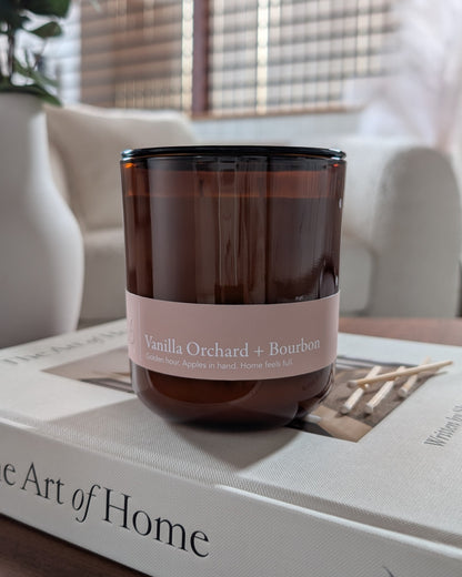 Vanilla Orchard + Bourbon scented candle in an amber vessel on top of a book in a cozy indoor setting. 