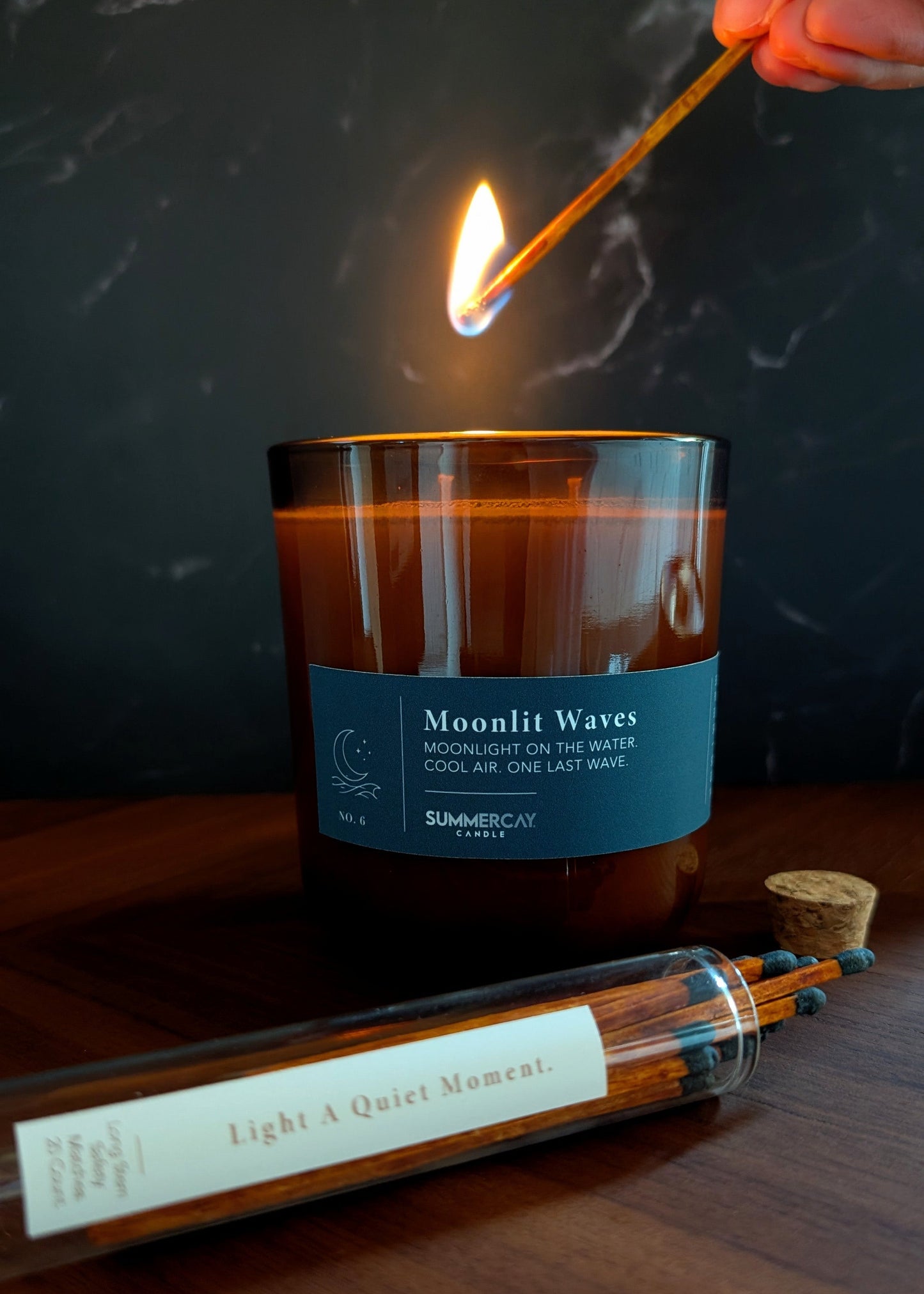 Candle labeled 'Moonlit Waves' being lit with a match on a wooden surface.