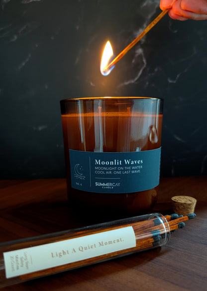Candle labeled 'Moonlit Waves' being lit with a match on a wooden surface.